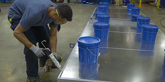 Video still of a worker using a caulk gun to seal a series of metal panels in a metal frame

