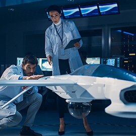 Two people in white lab coats looking at a small aircraft model in an office.