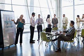 Standing woman explains charts to sitting man in office with other people standing and conversing.