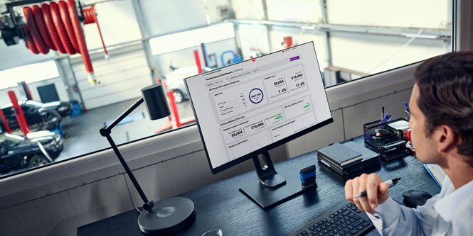 A man looks at a desktop computer screen. An autobody shop is shown in the background.
