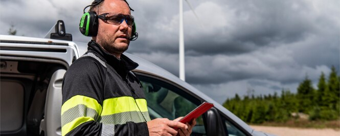 A person is standing outdoors near a vehicle, wearing a high-visibility jacket with yellow and gray stripes and green ear protection.
