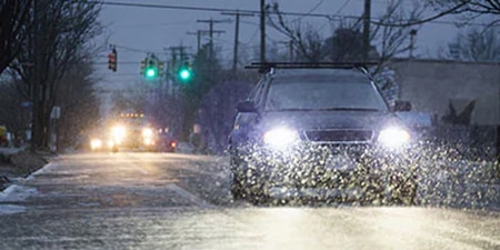 A car’s headlights illuminate falling snow on a road at dusk.
