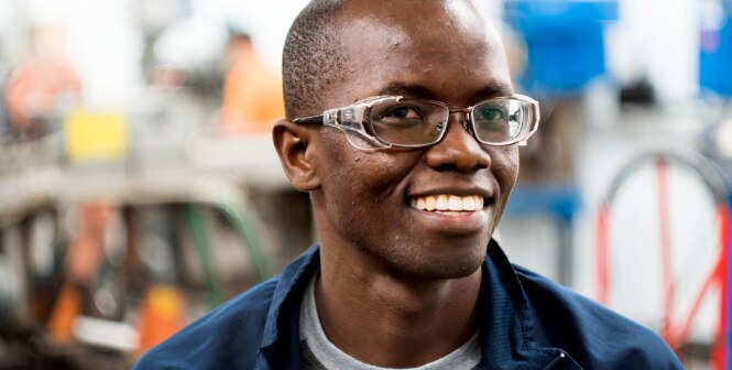 A black man is wearing glasses and dark jacket, standing in what appears to be a workshop or industrial setting.
