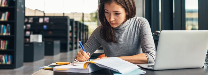 A woman is seated at a desk in a library, writing in a notebook with a pen.
