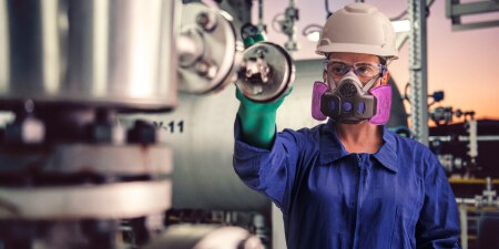 The image shows a person in protective gear&mdash;hard hat, gloves, respirator&mdash;working in an industrial setting, inspecting equipment amid machinery and piping.
