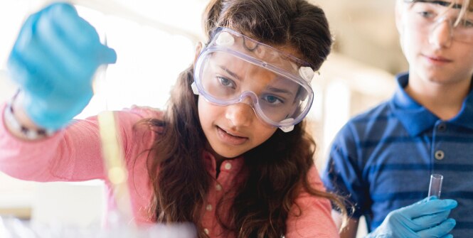 Lab workers in safety goggles and blue gloves handle a test tube while one person pours liquid during an experiment.
