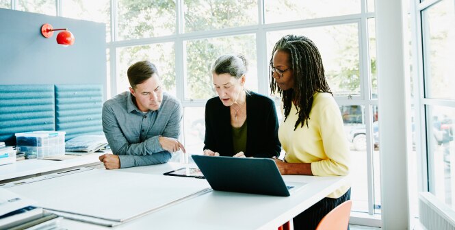 Three persons sitting around a table in a bright, modern office setting with a open laptop in front of them.
