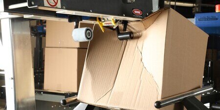 A cardboard box being crushed by an industrial packaging machine, illustrating structural stress.
