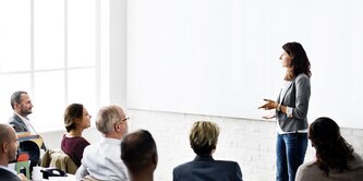 A woman leads a business presentation in front of a large white screen for an audience
