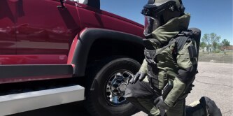 A person wearing a full bomb disposal suit is kneeling next to a red vehicle, inspecting it. The scene is outdoors on a sunny day.
