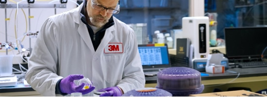 A 3M lab worker wearing safety glasses and a lab coat, inspecting a sample.
