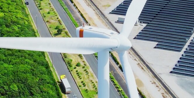 A large wind turbine is positioned near a canal bordered by lush green trees on one side and a field of solar panels on the other.
