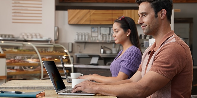 Two people sitting at a coffee shop sitting on their laptops with 3M Privacy film on them with cups of coffee sitting next to them.
