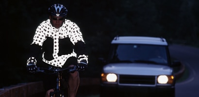 A bicyclist in a highly reflective jacket rides on a road in the dark in front of a car. 
