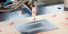 A robot arm applies adhesive to a metal plate in a rectangular pattern
