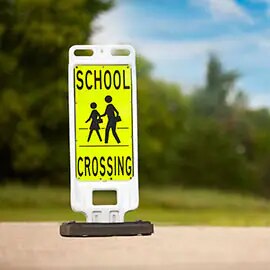 Standalone sign with yellow background and black image of two people crossing street and words “school crossing.”