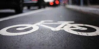 Close-up of biker symbol in white, on black pavement, in urban bike lane with cars in the background.

