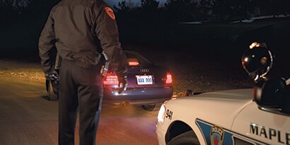A law enforcement officer faces a car whose rear license plate is illuminated by headlights in the dark.
