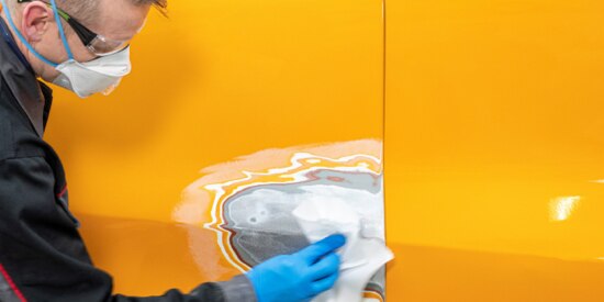 Man in a mask applying surface leveling compound to the side of a yellow vehicle.
