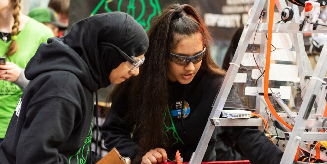 Two individuals wearing black hoodies are working together on a technical project involving a metal structure with wires and electronic components.
