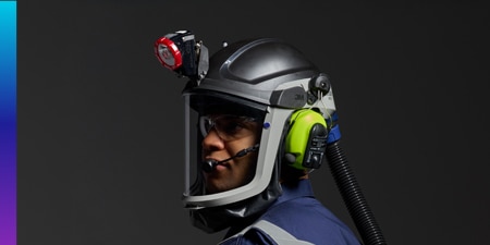 Person wearing a black protective helmet with a full-face shield, mounted light, PÈLTOR earmuffs, and powered air-purifying respirator hose.
