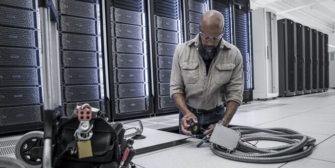 worker maintaining sub-floor electrical equipment in front of server racks
