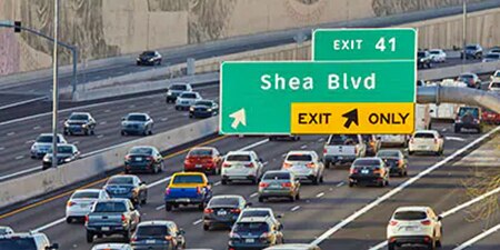 A large highway sign is highly visible over a busy highway.
