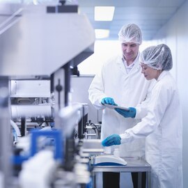 Two people in white lab coats and hair nets pointing at a small screen while they work.