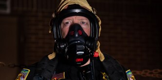 A person in a tactical uniform with a 3M gas mask, protective hood, and patches, including an American flag, in a dimly lit setting.
