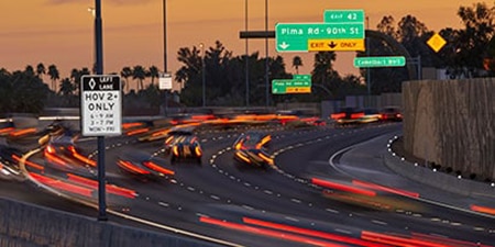 Cars speed past illuminated road signs on a highway at sunset.
