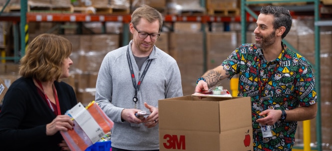 Three employees with blurred faces stand around a 3M-branded box in a warehouse, one sealing the box while another holds colorful folders.
