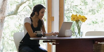 With trees in the background and flowers on the table, a woman types on a laptop computer.
