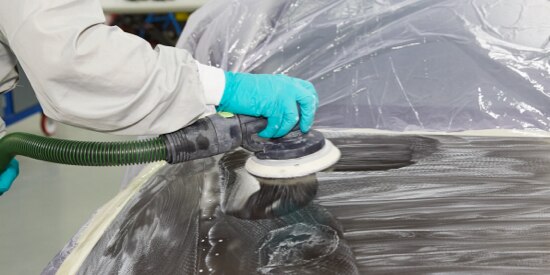 Technician in protective coveralls and gloves doing surface finishing on the hood of a vehicle.
