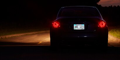 A car drives down a dark road at night with a highly visible, illuminated rear license plate.
