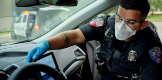 A police officer wearing a uniform and blue gloves is reaching into a car, interacting with a device on the dashboard. Another police vehicle is visible in the background.

