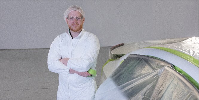 Image of a worker in white coveralls standing next to a vehicle masked off with tape and masking film
