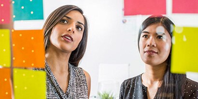 Two women assessing Post-it Notes that are posted to a glass pane.
