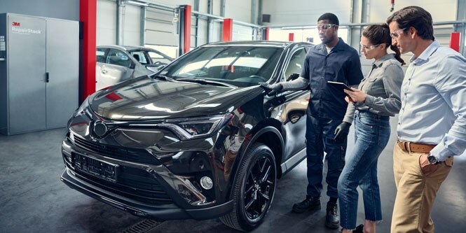 Three people in an autobody shop stand next to a vehicle, gesturing and talking. The person in the middle holds a tablet.
