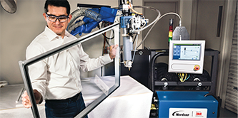 Image of a man holding a frame to which 3M™ VHB Extrudable Tape has been applied by the robot in the background
