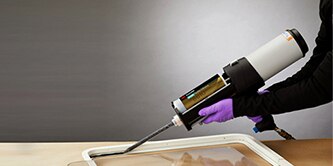 Image of a worker using a pneumatic applicator to apply adhesive to a frame in order to bond a piece of clear material
