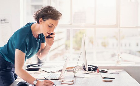 Woman on phone standing over desk taking notes in bright white sun-bathed office with large windows

