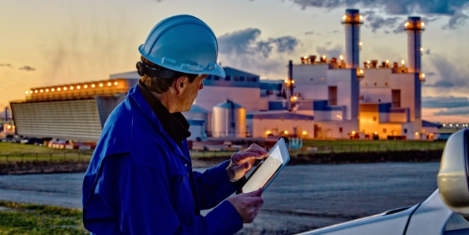 Worker with a hard hat choosing something on his iPad in front of a factory in the background.
