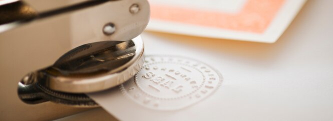A metal embossing tool is pressing a seal onto a piece of paper, creating a raised, circular imprint.
