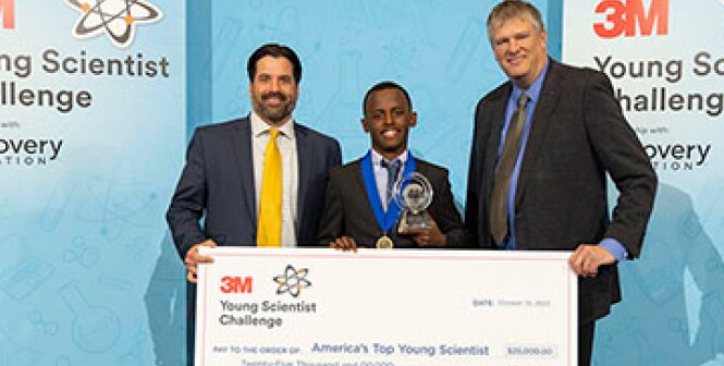 Three individuals are standing together holding a large ceremonial check for the 3M Young Scientist Challenge.
