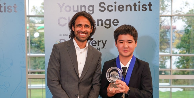 Two people stand at a Young Scientist Challenge backdrop, with one holding a clear trophy and wearing a medal.
