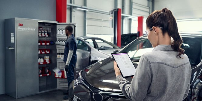 A woman looks at a tablet screen while standing in front of a vehicle in a body shop. In the background, a technician looks inside the 3M™ RepairStack™ cabinet.
