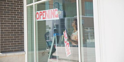 Two people hanging “opening soon” signs in window of commercial space.
