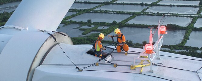 Two workers in safety gear are performing maintenance on a large wind turbine.
