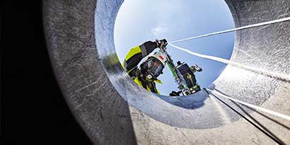 Worker in protective gear looking down into a confined space entry portal.
