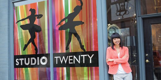 A woman leans against a storefront with large, bold window wraps displaying dancers and the words “studio twenty.”
